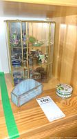Miniature brass and glass display case with assorted small trinket boxes inside, placed on wooden shelf.
