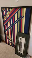 Large framed abstract geometric art print with diagonal stripes in red, gold, blue, and dark green leaning against wall next to smaller framed print.