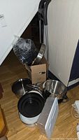 Photo showing assorted pots and frying pans along with a metal frame with black handles and a cardboard box.