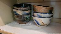 Stack of six stoneware bowls showing a variety of natural glaze colors and textures including dark green, black, blue, cream, and brown tones.