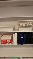 Seven binders and paper storage bins on shelves, showing arrangement and quantities of items.