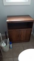 Wooden cabinet on wheels with shelf and doors shown in a corner of a room, next to bathroom accessories on the floor.