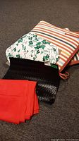 Two striped chair cushions, one reverse side floral pattern placemat, one brown woven placemat, and folded red plastic table cloth on carpeted floor.