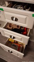 Three kitchen drawers partially open showing cutlery trays with forks, knives, and spoons, along with can opener, apple-shaped cover, and trash bags.