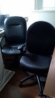 Two black office desk chairs side by side, one with fabric upholstery and one with faux leather upholstery, both equipped with wheels and adjustable height mechanisms.