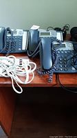 Three AT&T grey desk telephones with digital caller ID displays and coiled handsets on a wooden desk; white extension cords are visible.
