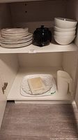 Stack of dinner plates, lunch plates, black tea pot, four white bowls, white milk jug, white trays and glass platter inside cabinet shelf