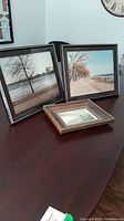 Three framed artworks on table: two larger square photographs of walkways by water and one smaller framed signed print with painted landscape.