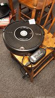 Black round iRobot Roomba Pet Series robot vacuum cleaner on wooden chair with charger adapter beside it.
