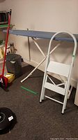 Photo showing Vileda broom with red handle, matching dustpan, gray kitchen garbage can, blue covered ironing board, and white two-step ladder.
