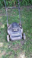 Black & Decker ElectroMulch electric lawn mower front view, showing top and parts of handle and wheels. The mower body is black with visible wear, placed on grass in outdoor setting.