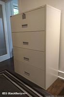 Photo showing beige rectangular metal file cabinet with four drawers, recessed handles, lock with key in top drawer, placed indoors near door and wall.