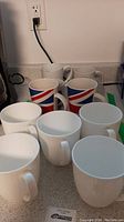 Photo showing eleven assorted mugs arranged on a kitchen counter, including nine plain white mugs and two mugs with a red, white, and blue striped pattern.