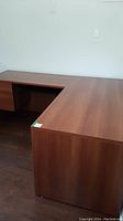 Photo of L-shaped office desk showing the front and part of the side with reddish-brown wood finish and clean surface.