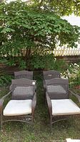 Front view of four brown resin wicker chairs with beige cushions arranged on lawn under tree