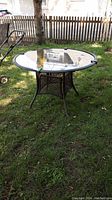 Full view of round patio table standing on grass in outdoor setting, showing glass top and metal/rattan-like base structure with four legs.