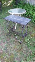 Photo showing round patio table with clear glass top and four legs next to rectangular slate-top accent table with wrought iron legs on grass.