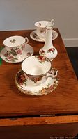 Photo showing three tea cups with matching saucers and one tall bud vase, all with floral patterns and gold trim.