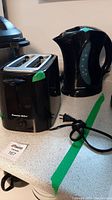 Photo showing Proctor Silex toaster and black electric kettle side by side on a kitchen counter with visible power cords and green tape.