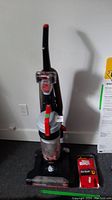 Upright Bissell PowerForce vacuum cleaner with black, gray, and red accents, standing on carpeted floor with a few accessories attached.