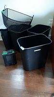 Photo showing four black wastebaskets of varying sizes, one black metal wire walker basket, and one Honeywell HZ-318C heater placed on a wood floor near a wall outlet.