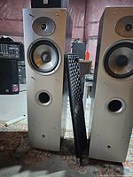 Pair of Athena Technologies floor standing speakers with one grille removed and leaning against a speaker, showing the drivers and bass reflex port on a silver front panel.
