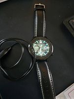 Front angled view of black smart watch on black surface with charger cable coiled next to it, showing analog style watch face with 12,3,6,9 hour marks