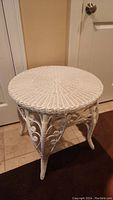White antique wicker table with woven round tabletop and decorative scroll woven wicker design on skirt and legs photographed against beige wall and tiled floor.