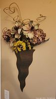 Front view of a decorative metal wall vase with artificial flowers including hydrangea and magnolia-like blooms and yellow accent flowers.