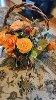 Woven basket filled with artificial orange roses and fall-themed greenery on a patterned carpet.