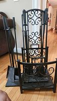 Complete fireplace tool set with poker, broom, shovel in stand and decorative firewood holder and screen, all black metal with scrollwork.
