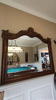 Front view of the large hand-carved wooden framed antique mirror mounted on a beige wall above white paneling, reflecting a room with a billiards table and stained glass hanging light fixture.