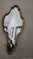 Photo showing the ornate shaped mirror hung on a wall with a gold-toned aged finish reflecting part of a room.