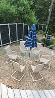 Top angled photo showing hexagonal glass table with umbrella and six beige cushioned chairs surrounding it, set outdoors on gravel circle.