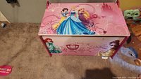 Pink Disney Princess-themed storage chest front and top views, featuring Cinderella, Rapunzel, Snow White, Ariel, and Belle artwork.