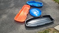 Four varied snow sleds on grass: orange and black large flat sleds, blue saucer sled, and blue child seat sled.