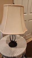 Table lamp with cream fabric shade on a white wicker table next to a white door and beige carpet