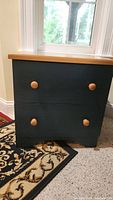 Photo of small two-drawer wooden end table with dark blue painted body and natural wood top and knobs beside window on patterned rug