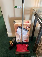 Red Dirt Devil Broom Vac with silver handle and black bristles standing upright next to its box featuring product image and branding.