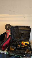 Photo showing Milwaukee angle grinder with orange body and black disc, inside a red carrying bag, next to Dewalt drill case.