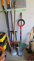 Full view of spades, shovels, and digging tools stacked against wall with some handles visible