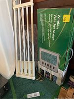 DSE Wireless Weather Station in green and white box standing upright, with adjacent white curtain rods leaning against the wall.