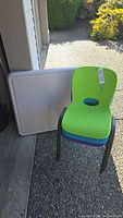 View of a green stackable kids chair leaning against a folded beige plastic top kids table showing the plastic back and steel frame chair design.