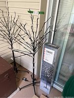 Two assembled Noma Canvas light up blossom trees with stands, plugged in, next to one unopened box showing the product image and brand.