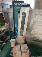 Full view of the white NOMA tower fan standing upright near its original box, with remote and accessories wrapped in plastic placed nearby.