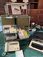 Photo shows metal rolling office cart with shelves holding green hanging files, Sharp calculator, Brother fax machine, Underwood electric typewriter in case, 3-hole punch, and various small office supplies in boxes and containers.