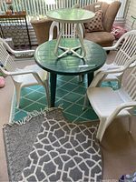 Photo showing green round plastic patio table with four white plastic chairs and a small round glass top side table with white metal legs, all on a green patio rug with diamond pattern.