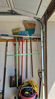 Full view of rakes, brooms hanging on wall with shop vacuum and yellow extension cord below.