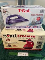 Photo of both boxed items: T-Fal Ultraglide Pro 1700W steam iron and Scunci 1000W pressurized steam cleaner stacked on green surface.