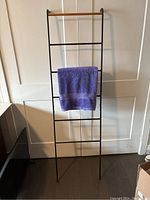 Full view of assembled black metal and wood ladder with one purple towel hanging on middle rung, showing entire length against white door backdrop.